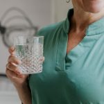 A happy middle-aged woman in a green shirt holds a stylish glass of water, smiling indoors.