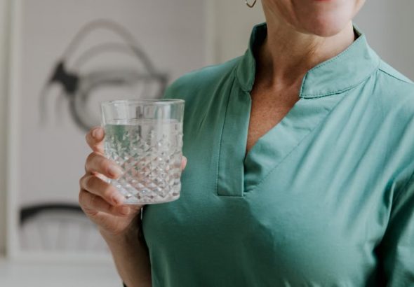 A happy middle-aged woman in a green shirt holds a stylish glass of water, smiling indoors.