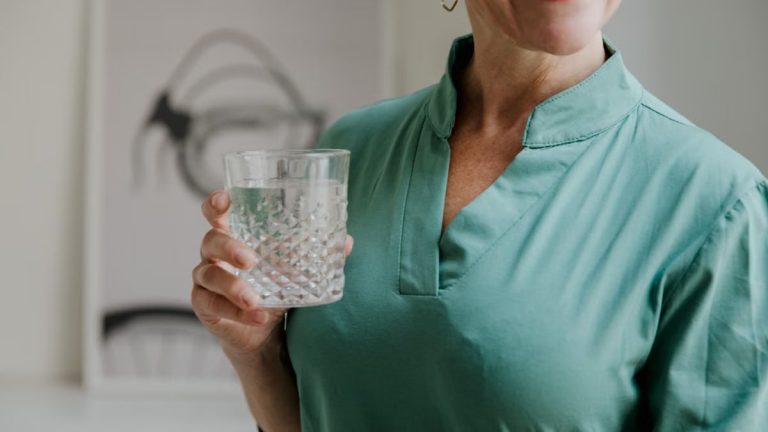 A happy middle-aged woman in a green shirt holds a stylish glass of water, smiling indoors.